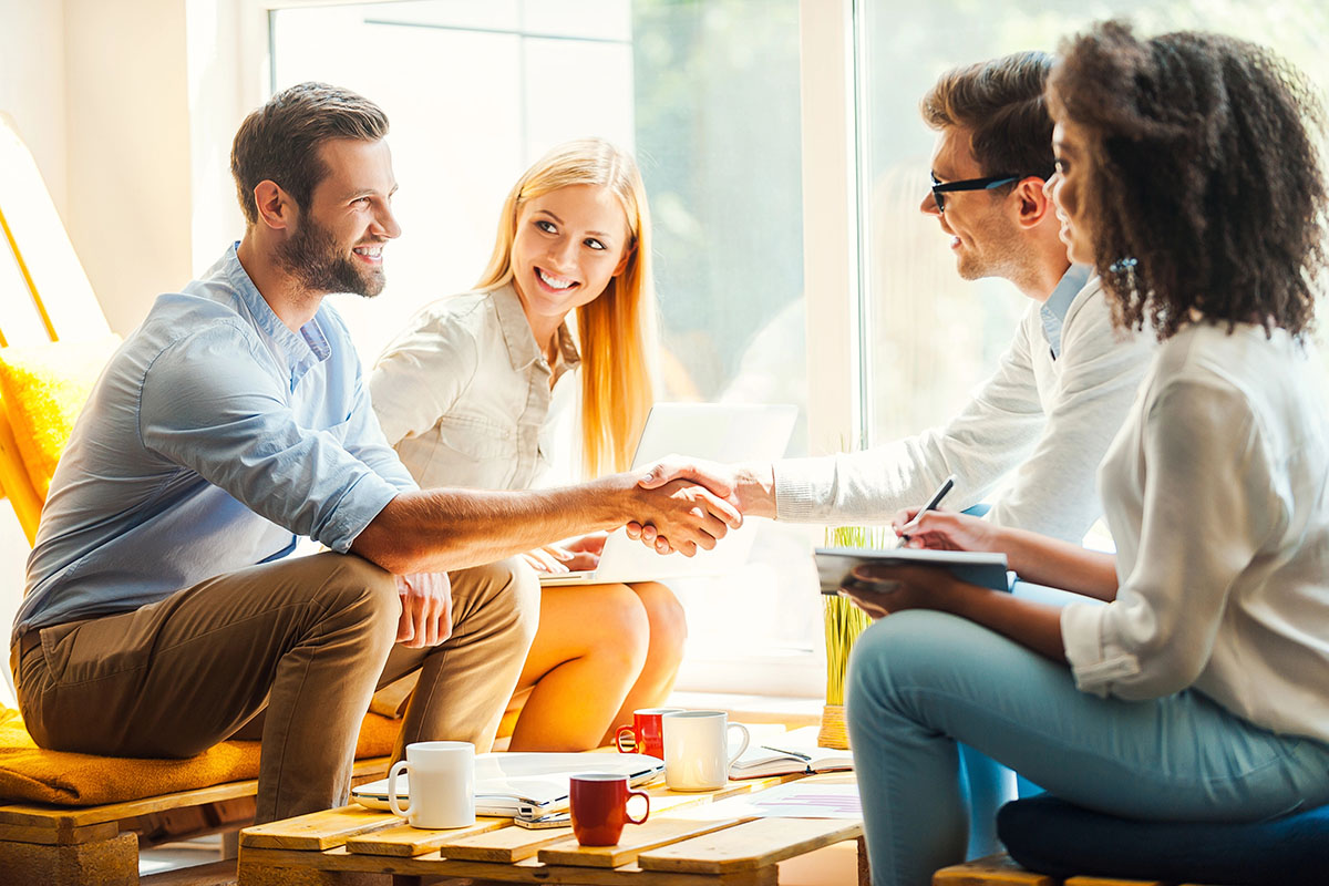 Two men are shaking hands over a coffee table in a business meeting.