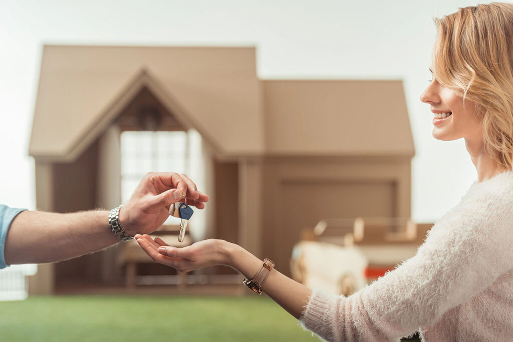 A smiling woman receives a set of keys from a man's hand, with a model house in the background.