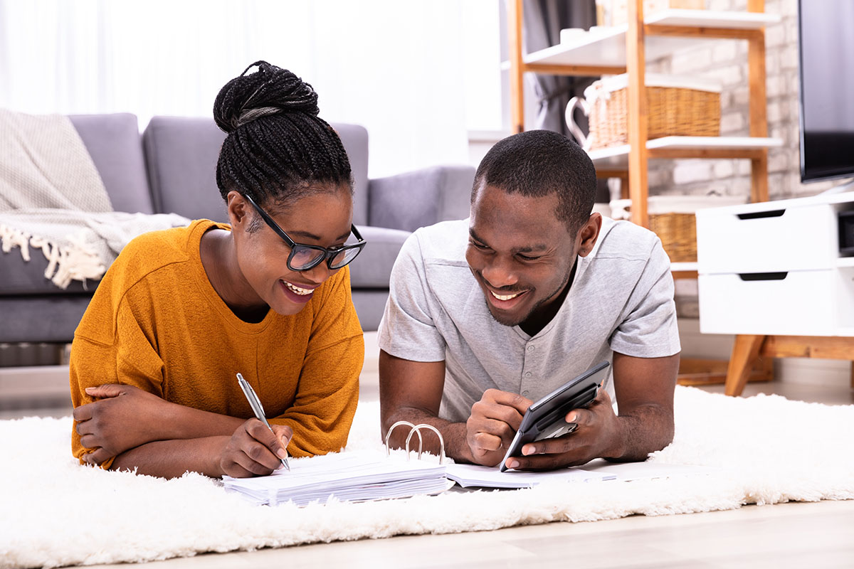 A young couple is lying on a fluffy white rug, engaged in study or work. The woman is smiling while taking notes with a pen, and the man, also smiling, is using a calculator. A cozy living room setting is visible in the background.