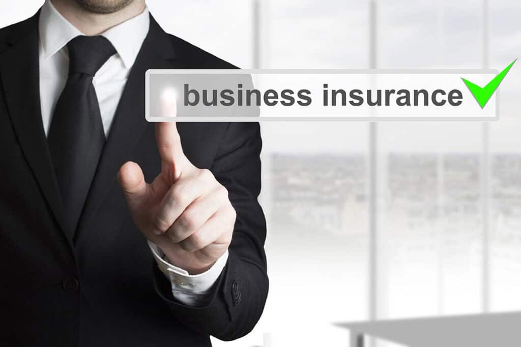A businessman in a black suit points at a translucent sign that reads "business insurance" with a green checkmark, set against a bright, blurred office background.