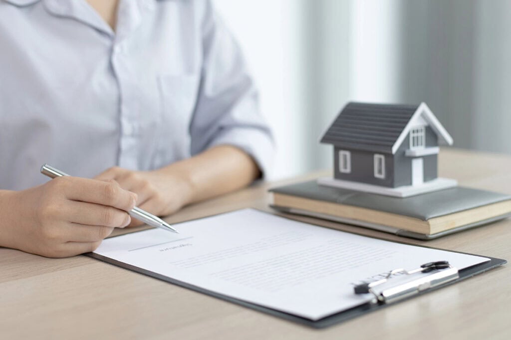A person’s hand holding a silver pen above a document, with a miniature house model and a notebook placed on a wooden table.