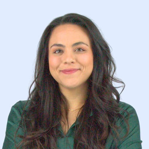 A smiling woman with long, wavy dark hair, wearing a green blouse, stands against a light blue background.