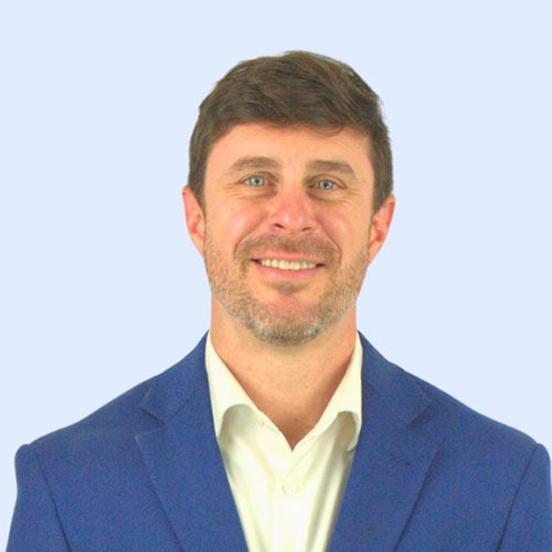 A smiling man with short dark hair and a beard, wearing a blue suit jacket and white shirt, against a light blue background.