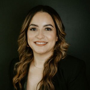A woman with long, wavy hair smiles confidently, wearing a black blazer against a dark background.