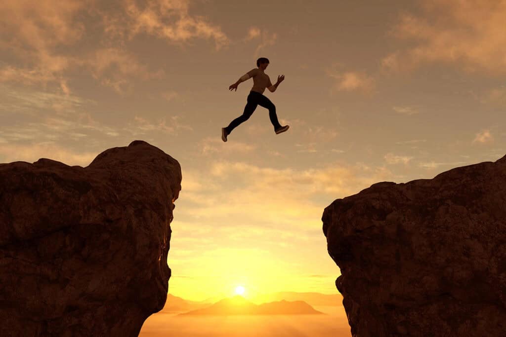 A silhouette of a person leaping across a gap between two rocky cliffs, against a vibrant sunset sky.