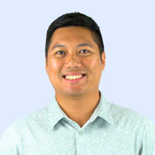 A smiling man with short black hair wearing a light-colored patterned button-up shirt, set against a light blue background.