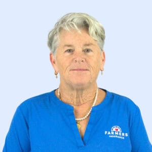 A middle-aged woman with short gray hair, wearing a blue shirt with the Farmers Insurance logo, stands against a light blue background, looking directly at the camera.
