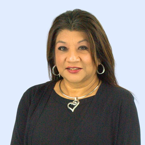 A woman with long dark hair, wearing a black blouse and a silver heart-shaped necklace, stands against a light blue background, smiling slightly.