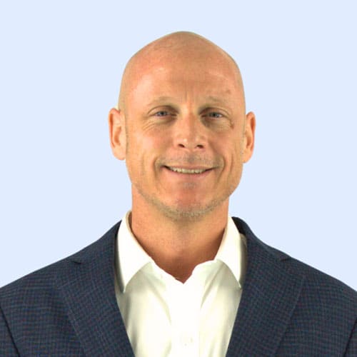A smiling man with a shaved head, wearing a dark blazer over a white shirt, poses against a light blue background.