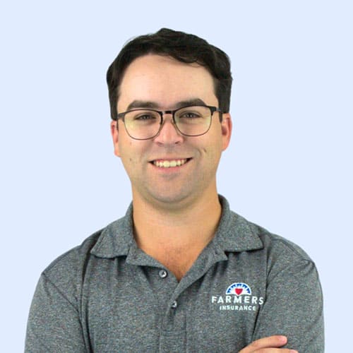 A smiling man with glasses stands with his arms crossed, wearing a gray polo shirt featuring the Farmers Insurance logo, against a light blue background.