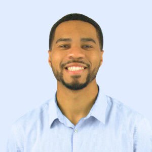 A smiling young man with a beard, wearing a light blue checkered shirt, against a solid light blue background.