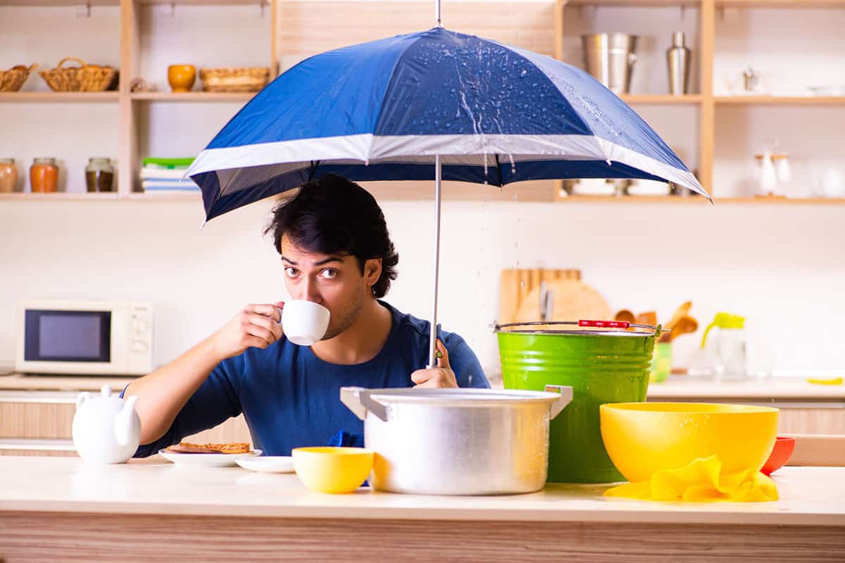 A man sits at a kitchen table, holding a white cup while looking at the camera. Above him, a blue umbrella catches dripping water from a leak. Nearby, there are various kitchen items, including a silver pot, a green bucket, and a yellow bowl. A plate of food and a teapot are placed on the table.