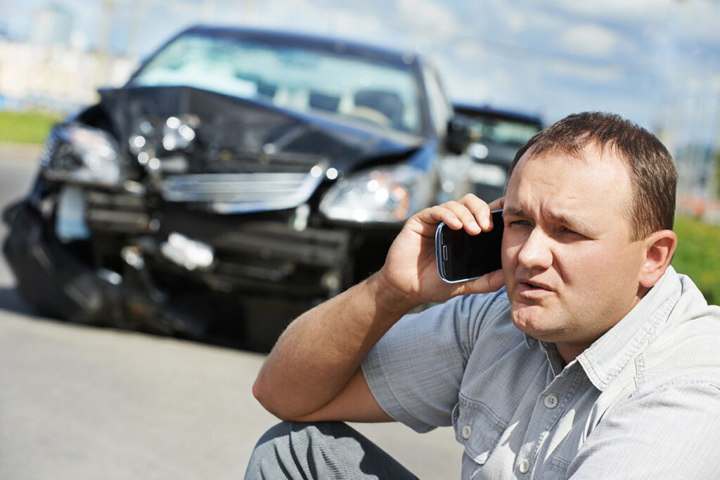 A man in a gray shirt sits on the ground, speaking on a phone, with a severely damaged black car in the background.