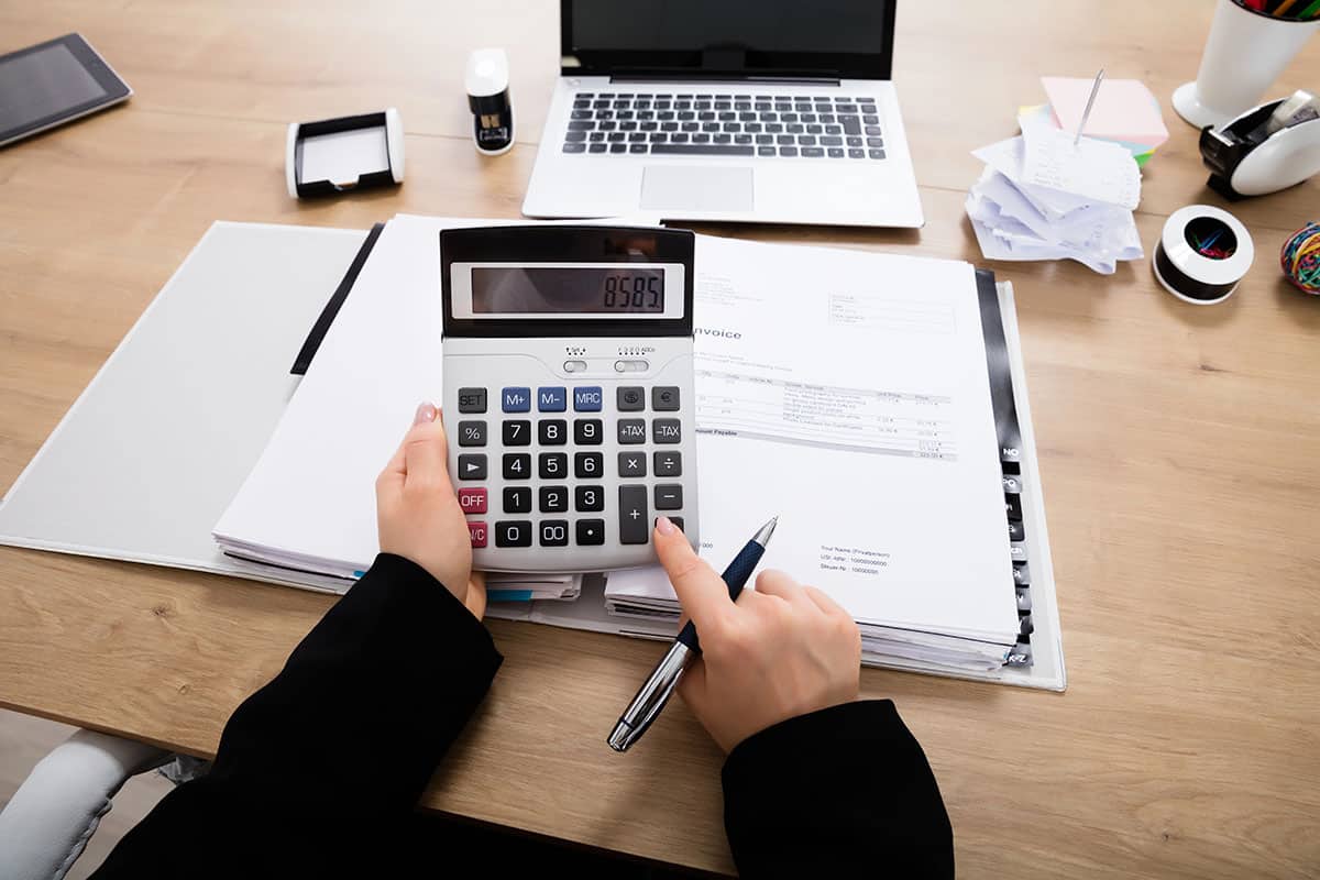 A person in a black jacket holds a calculator displaying the number 8585, while pointing at financial documents and using a pen on a wooden desk with a laptop and office supplies in the background.