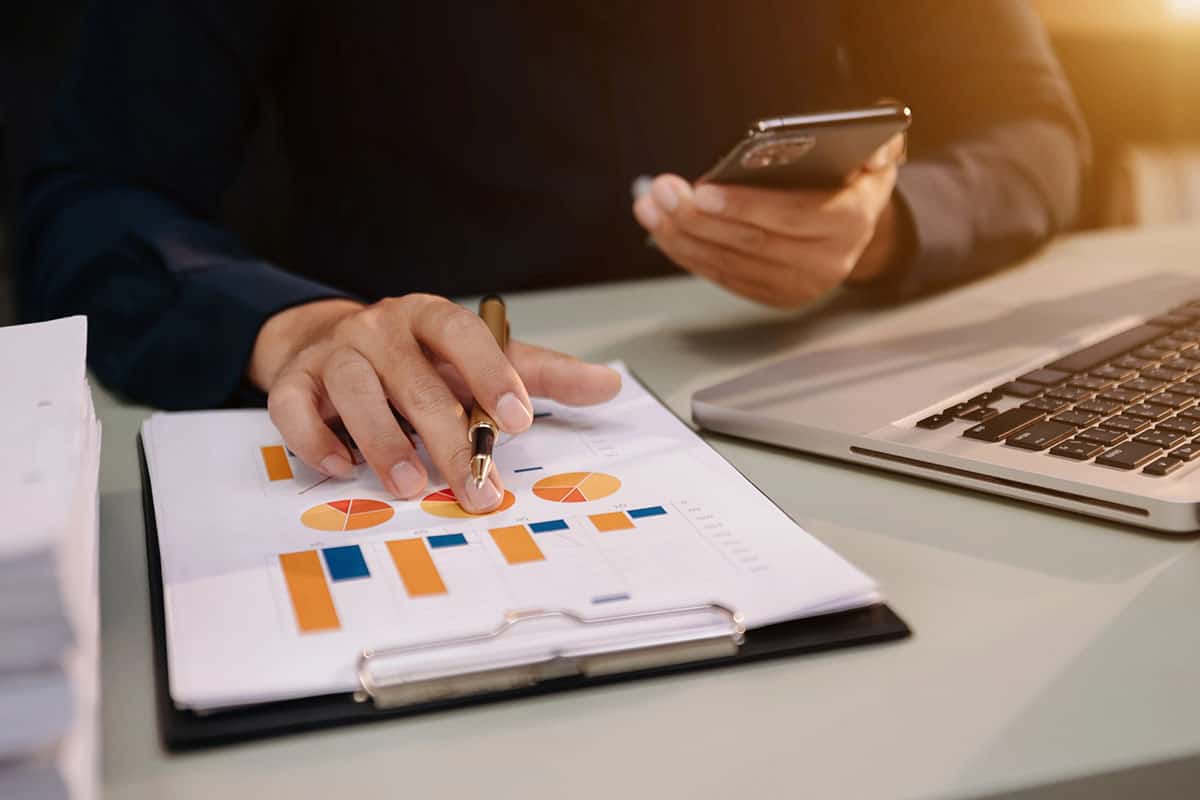 A person's hand holding a pen points to charts and graphs on a clipboard, while the other hand holds a smartphone. A laptop is visible on the desk along with stacks of paperwork.