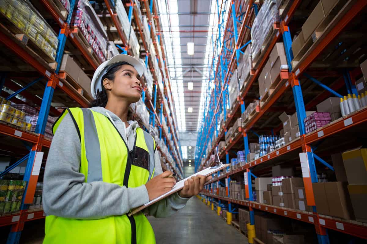 A woman wearing a hard hat and a high-visibility vest stands in a warehouse, holding a clipboard and looking upwards among tall shelves filled with boxes and products.