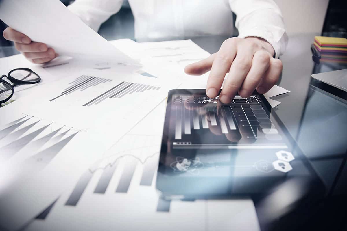 A person’s hand interacts with a tablet displaying digital graphs and charts, while other papers with financial data are scattered on a desk alongside a pair of glasses.
