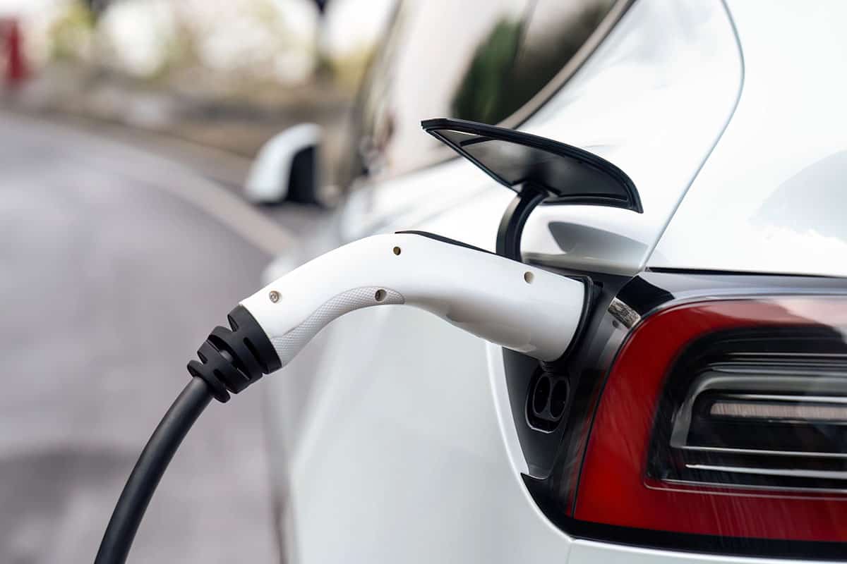 A close-up of an electric vehicle charging connector plugged into the charging port of a white car, with the vehicle's rear and road in the background.