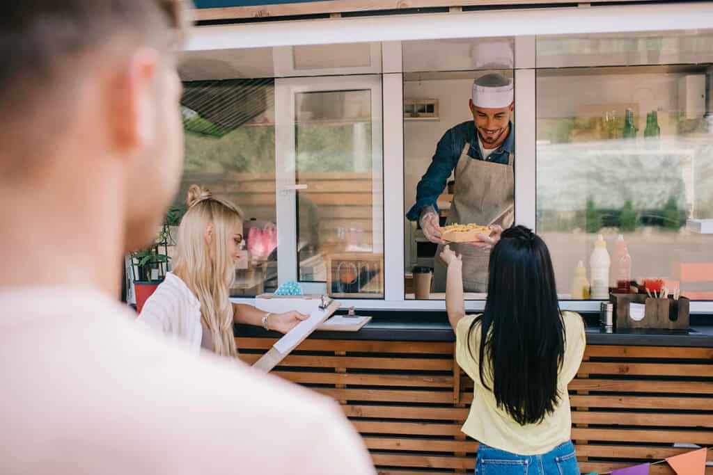 A smiling vendor in an apron hands a plate of food to a woman reaching through a service window, while another woman with a clipboard observes nearby.