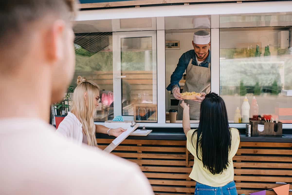 A smiling vendor in an apron hands a plate of food to a woman reaching through a service window, while another woman with a clipboard observes nearby.