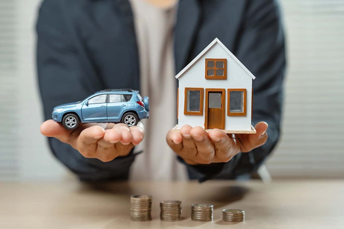 A person holds a small model car in one hand and a small model house in the other, with stacks of coins visible on a table in front of them.