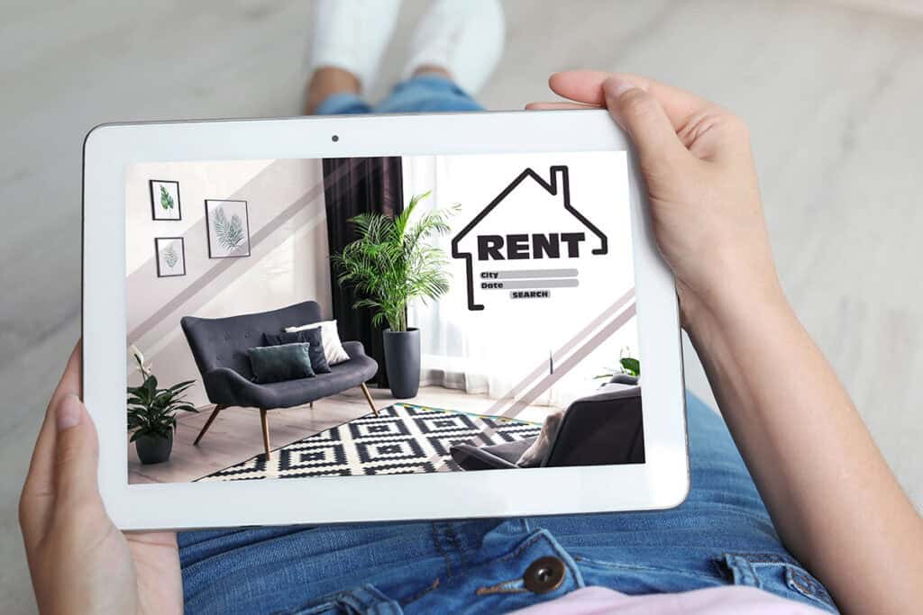 A person holds a tablet displaying a room for rent, featuring a stylish chair, decorative plants, and artwork on the wall. The tablet screen includes a "RENT" label and fields for city, date, and search options.