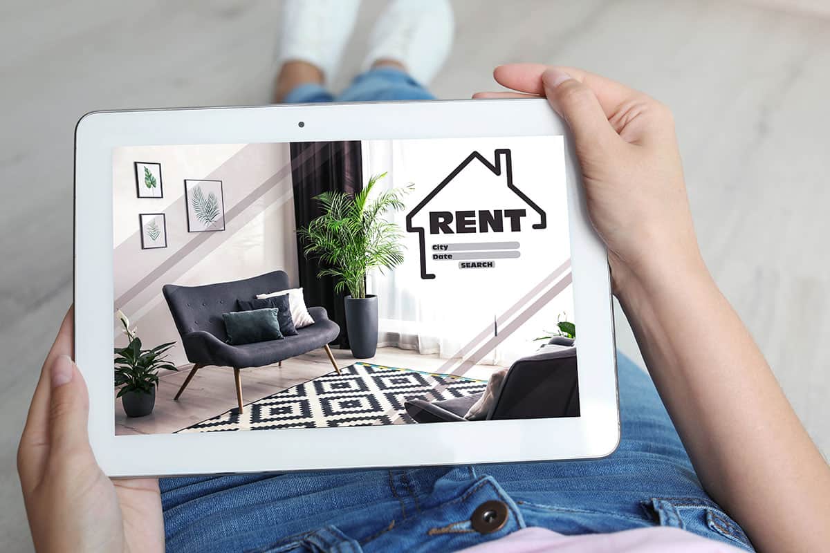 A person holds a tablet displaying a room for rent, featuring a stylish chair, decorative plants, and artwork on the wall. The tablet screen includes a "RENT" label and fields for city, date, and search options.