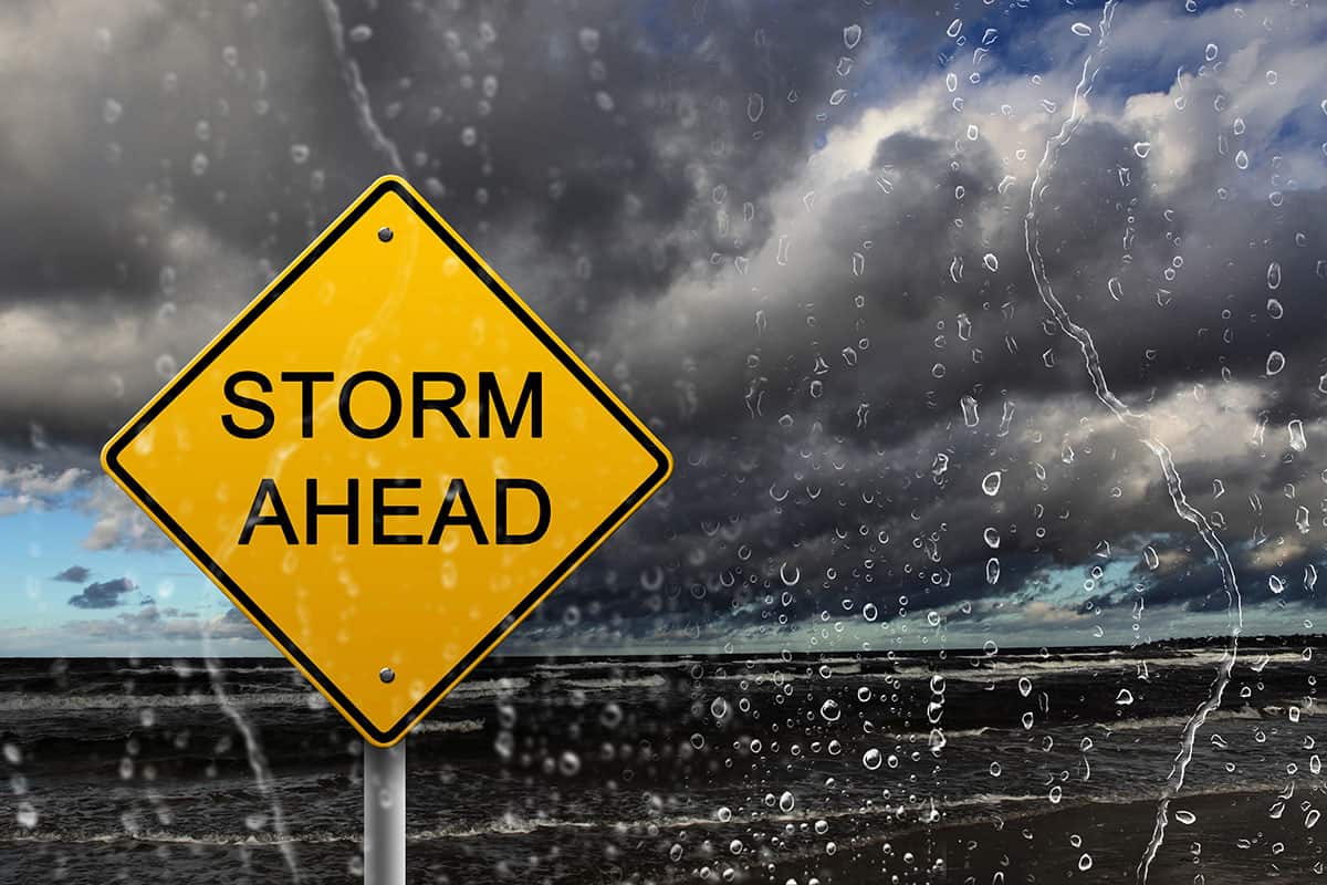 A yellow diamond warning sign reading "STORM AHEAD" positioned against a backdrop of dark, stormy storm clouds over a body of water, viewed through a rain-streaked window pane.