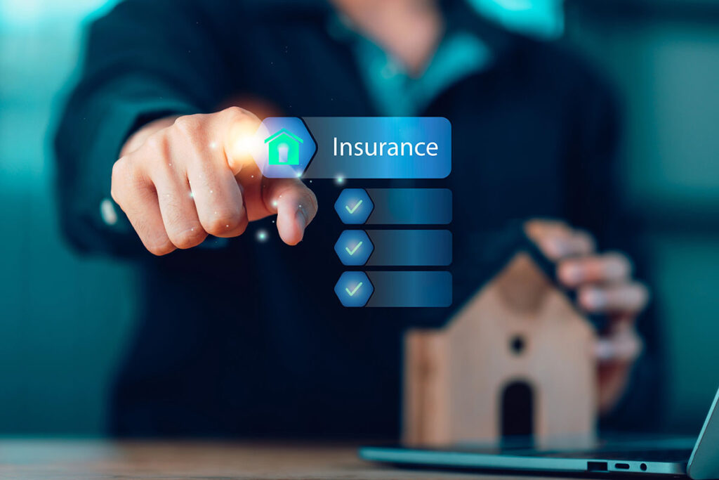 A person pointing toward a glowing digital interface button labeled "Insurance" with a house icon and a checkmark list, with a small model house sitting on a desk in the foreground.
