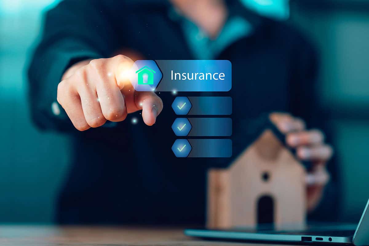 A person pointing toward a glowing digital interface button labeled "Insurance" with a house icon and a checkmark list, with a small model house sitting on a desk in the foreground.