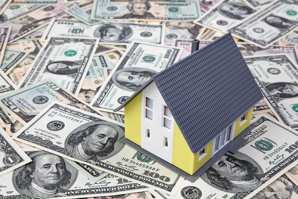 A small yellow model house with a grey roof resting on a bed of hundred-dollar and twenty-dollar U.S. cash bills, representing New Braunfels home insurance.