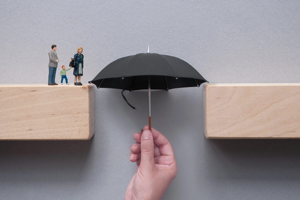 A hand holds a small black umbrella upward to span the gap between two wooden blocks, bridging the path for miniature figurines of a family.