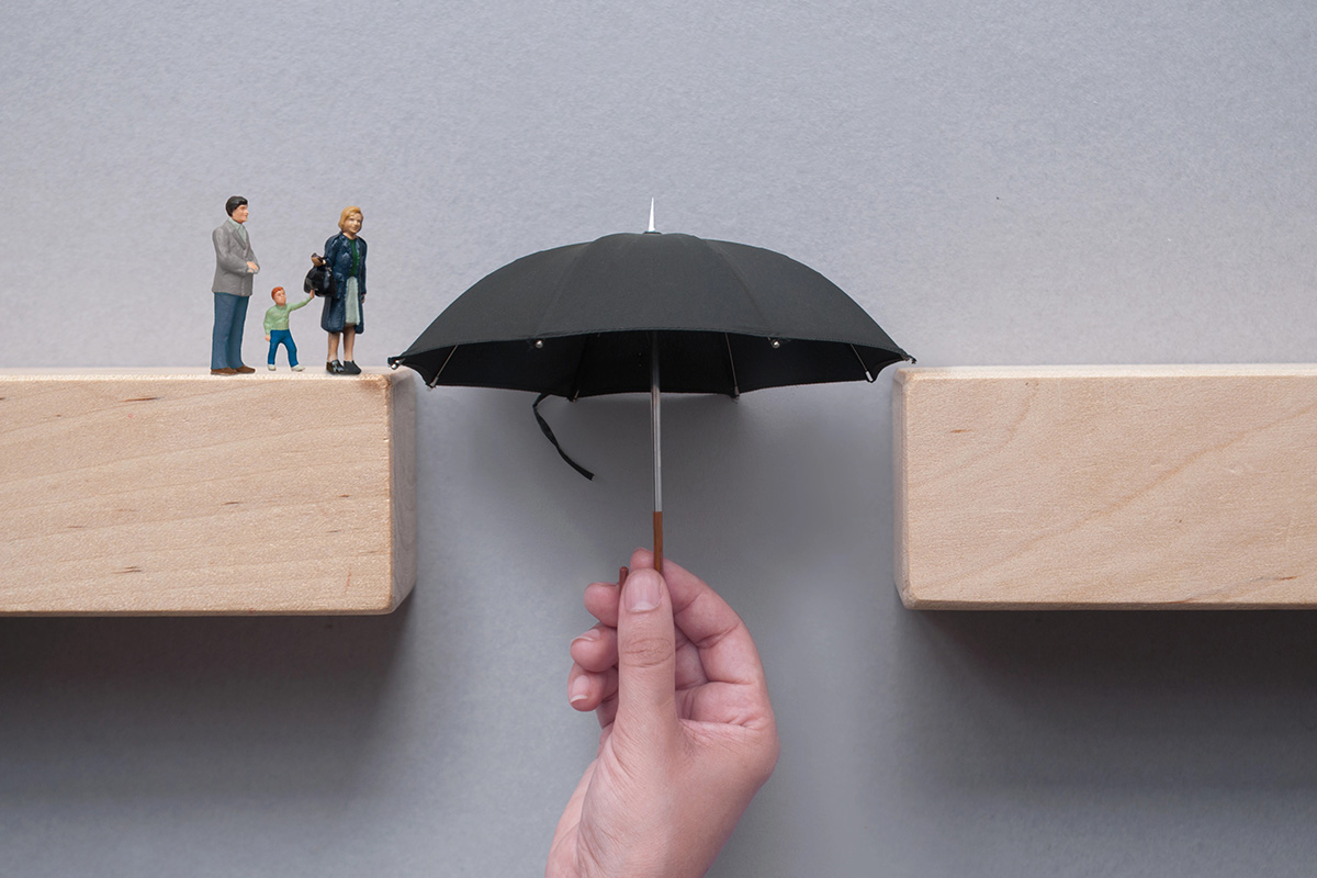 A hand holds a small black umbrella upward to span the gap between two wooden blocks, bridging the path for miniature figurines of a family.