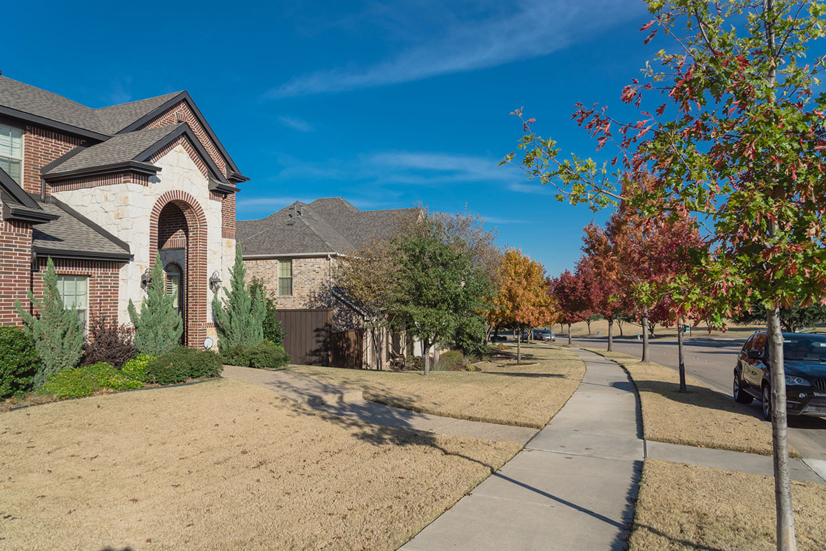 A residential suburban street featuring two-story brick and stone houses, manicured front lawns, and colorful autumn trees along a concrete sidewalk.
